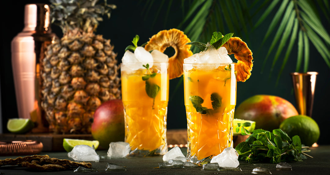Two tropical orange colored cocktails, with cocktail shaker, mangos, and a pineapple.