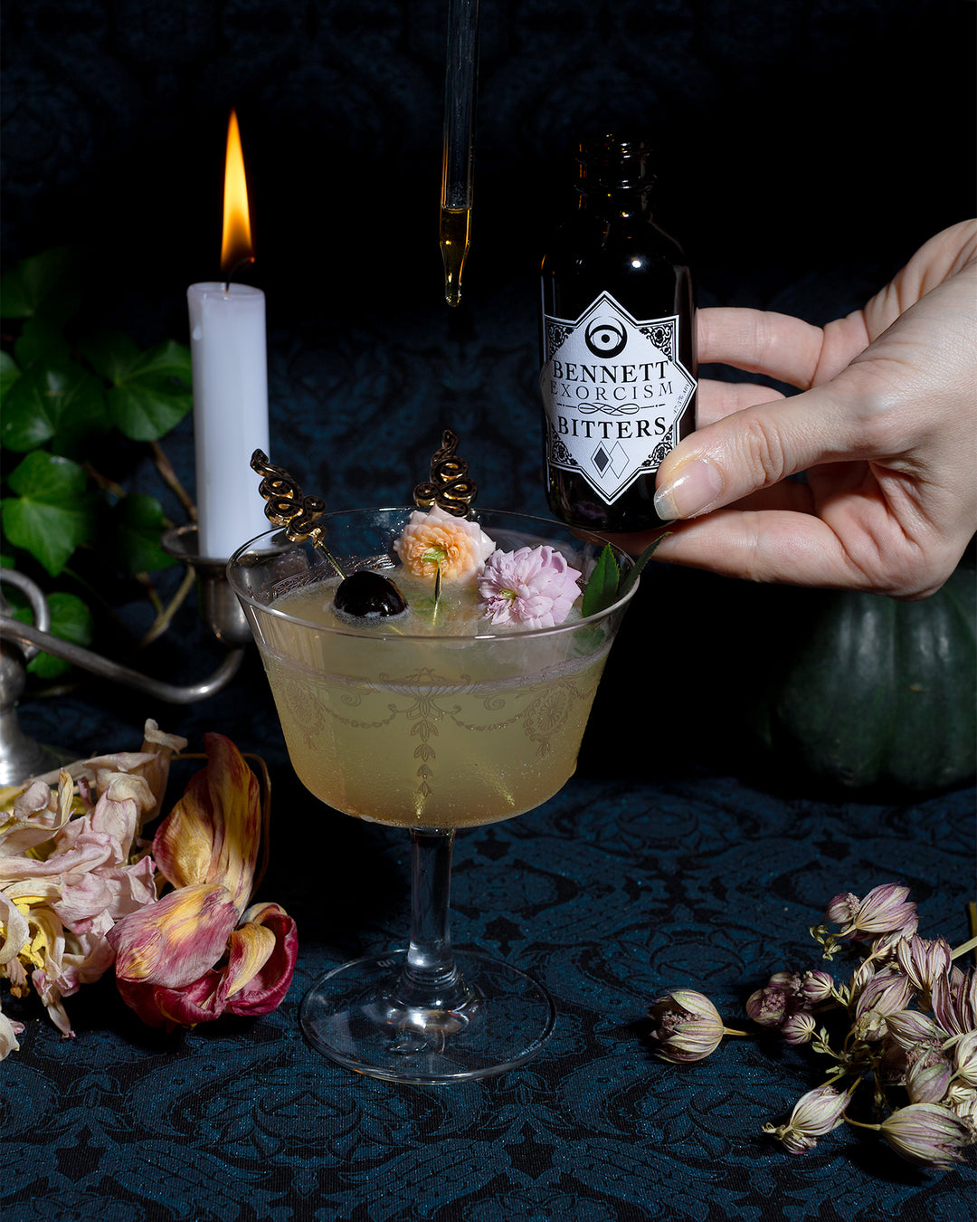 A hand dripping Exorcism Bitters into a Corpse Reviver number 2 cocktail in a moody setting with candles, a dark green pumpkin, and botanicals.