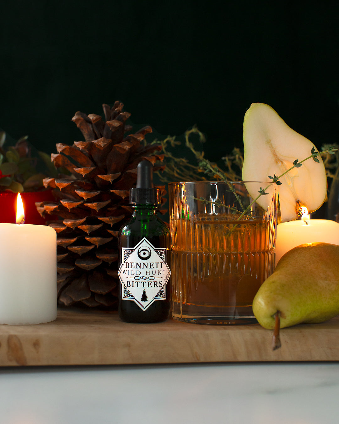 A bottle of Bennett Wild Hunt Bitters next to The Capricorn Maple Old Fashioned in a rocks glass with pear slice and thyme garnish, next to a pear, a large pinecone, and a glowing candle.