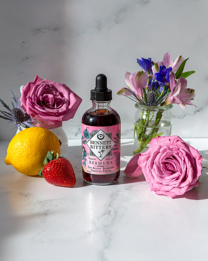 A bottle of tropical Bennett Bermuda Bitters with lemon, strawberries, flowers, and purple roses on a white marble background.