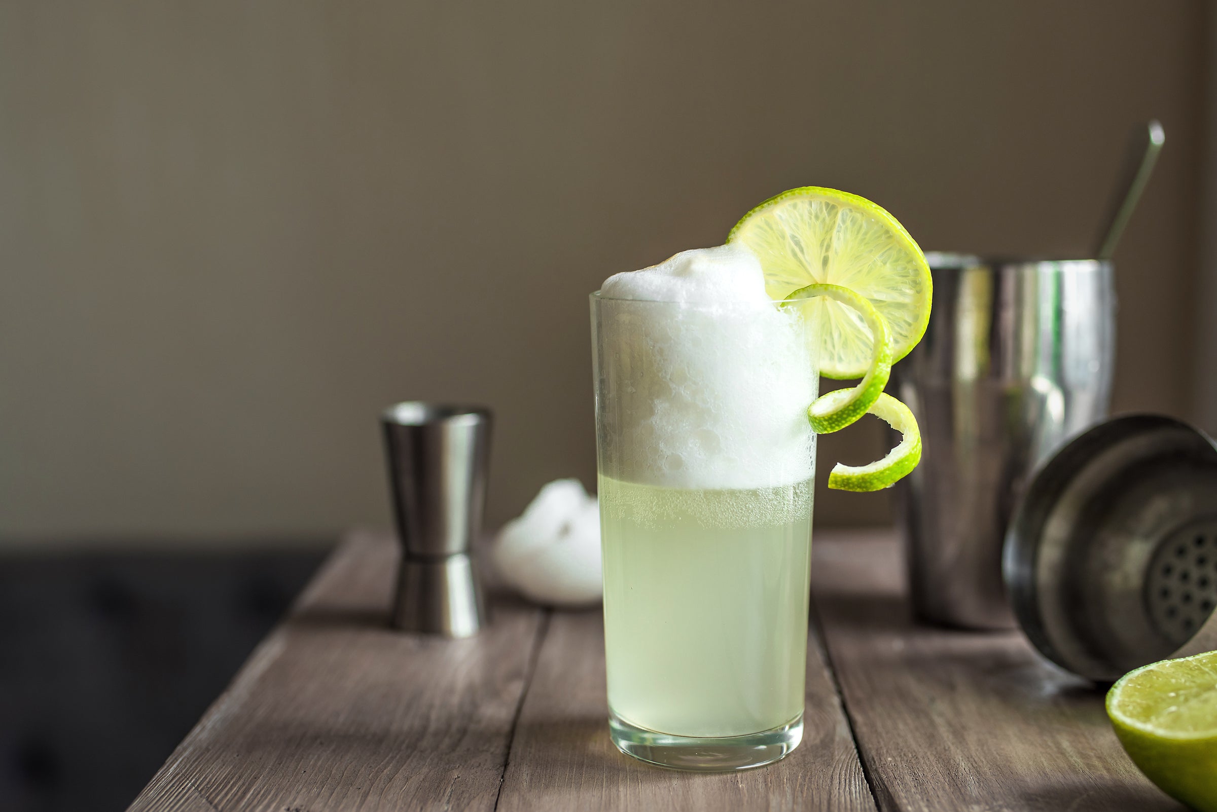A pale yellow gin fizz cocktail that has a nice frothy head with lime wheel and twist garnish, with steel cocktail tools in the background, on a grey wood table.