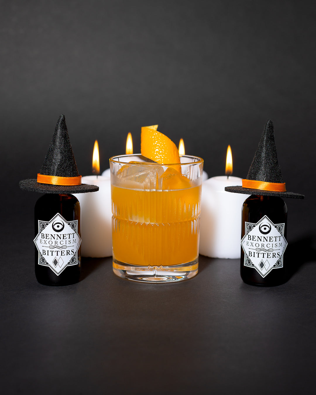 Glowing orange Witch Hat cocktail in a rocks glass next to bottles of Exorcism Bitters that have little witch hats on them, with candles in the background.