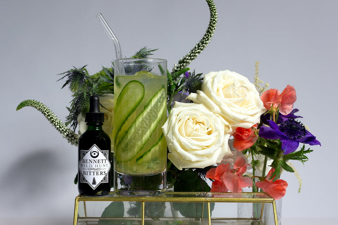 American Psycho cucumber gin buck next to a bottle of Bennett Wild Hunt Bitters, on top of a glass case and in front of white roses and various purple and white flowers.