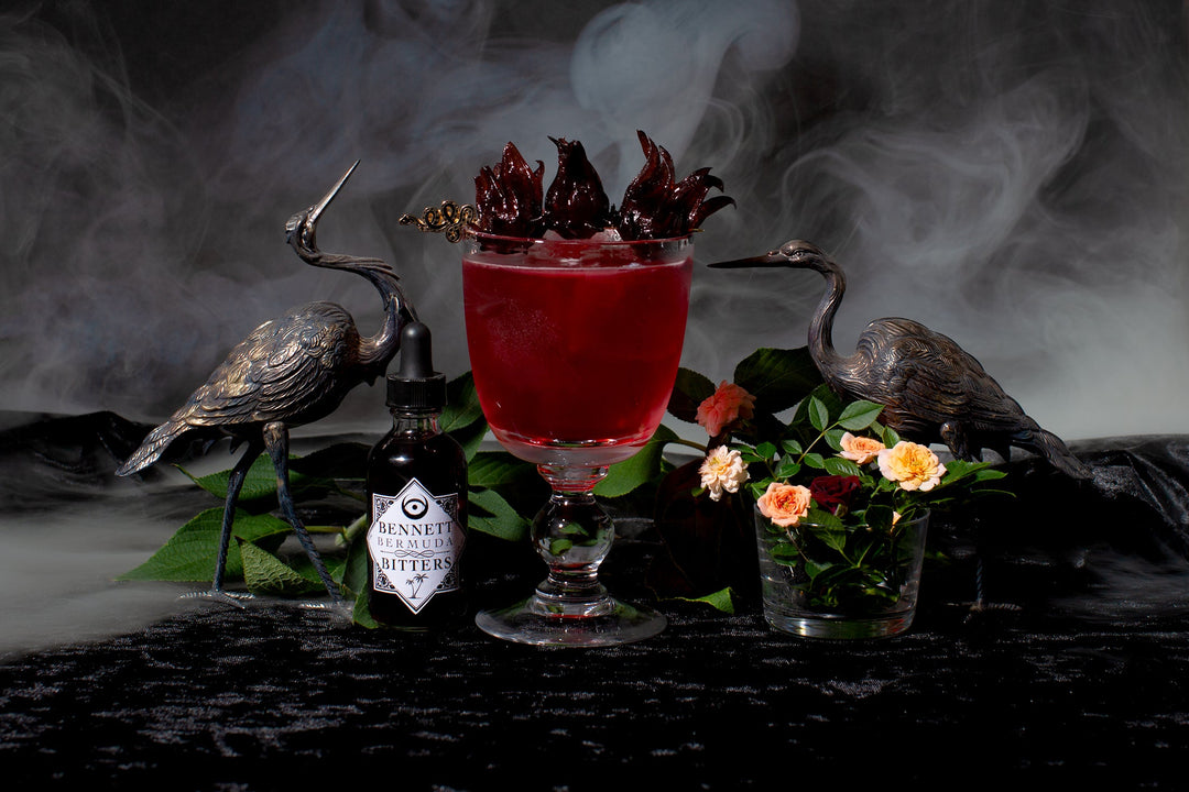 Bad Blood cocktail next to a bottle of Bermuda Bitters, two silver crane statuettes, and flowers; on black velvet; surrounded by smoke.