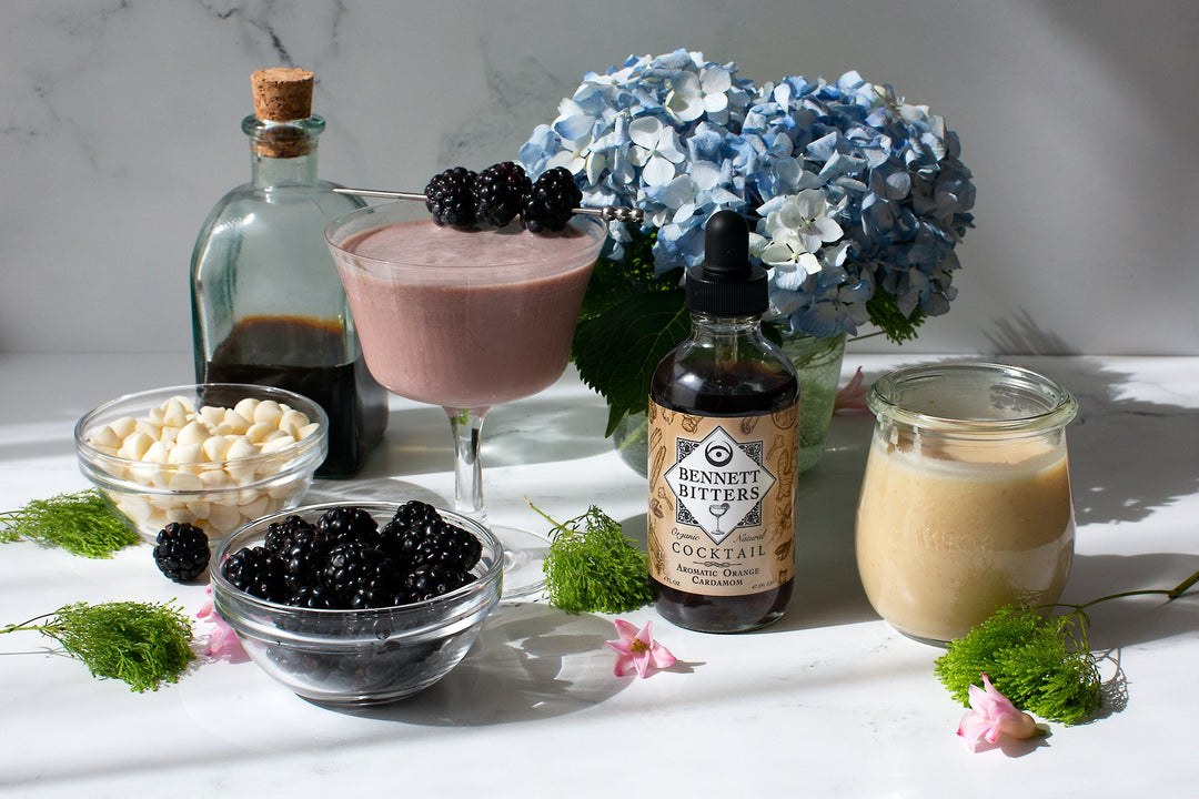 Midnight Sprite blackberry white chocolate espresso martini cocktail with blackberry garnish, in front of hydrangea flowers,  next to a bottle of Bennett Cocktail bitters, bowls of white chocolate and blackberries, and white chocolate sauce.