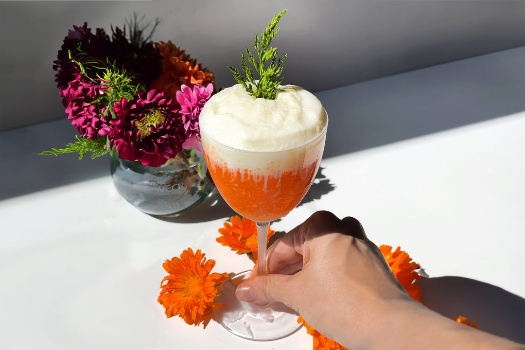 A hand holding The Carrot Cake cocktail, a bright orange drink in a Nick and Nora glass topped with white fluffy cream cheese foam, with orange and purple flowers around it.