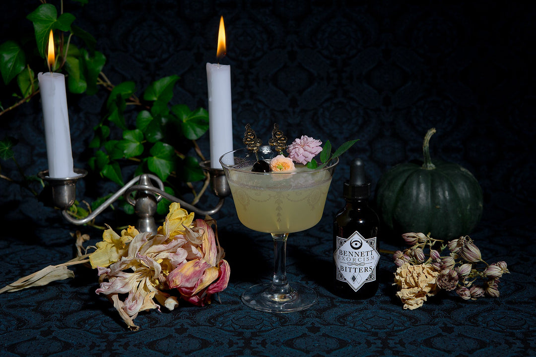 Bottle of Exorcism Bitters next to Corpse Reviver Number 2  cocktail in a moody setting with candles, a dark green pumpkin, and botanicals.