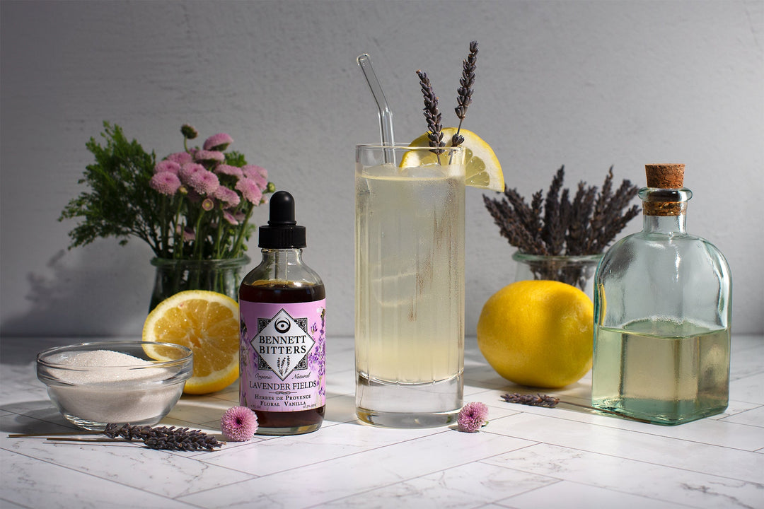 A Lavender Lemonade in a collins glass garnished with  lavender sprigs and a lemon wheel surrounded by Lavender Fields Bitters, flowers, lemons, and sugar.