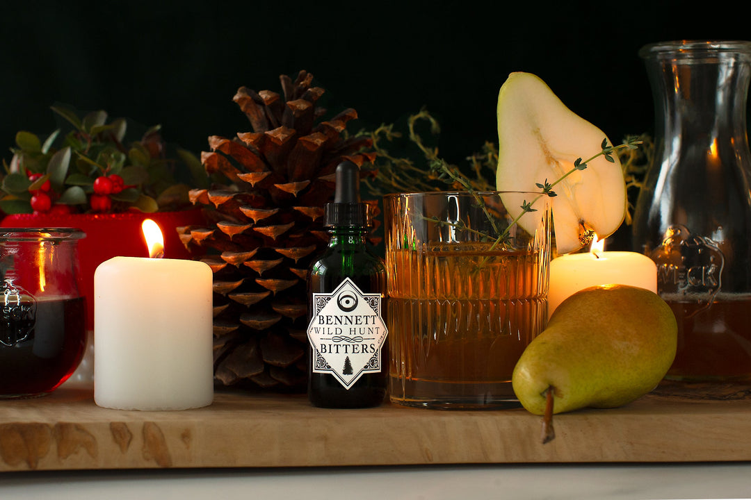 The Capricorn Maple Old Fashioned cocktail next to a bottle of Bennett Wild Hunt Bitters, glowing candles, a pear, a large pinecone, a wintergreen plant, thyme, and jars with liquid ingredients.