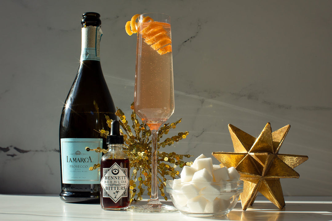 Pink Champagne Cocktail served in a Champagne flute with a lemon zest garnish in a white marble setting, surrounded by a bottle of Bermuda Bitters,  a bottle of Prosecco,  a bowl of sugar cubes, and New Years decorations.