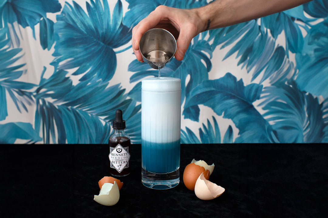 Hand pouring soda water into a blue gin fizz cocktail with a tall foamy head, surrounded by egg shells and a bottle of Bennett Bermuda Bitters.