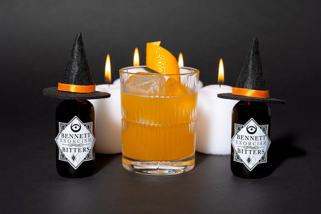 Glowing orange Witch Hat cocktail in a rocks glass next to bottles of Exorcism Bitters that have little witch hats on them, with candles in the background.