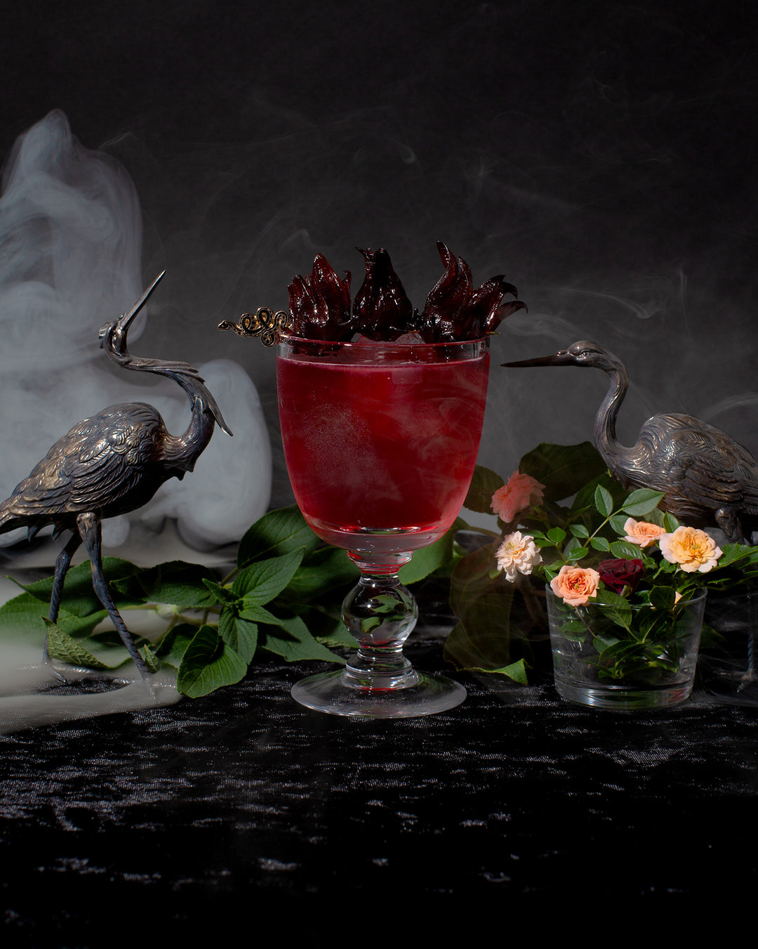 Bad Blood cocktail next to two silver crane statuettes, with fresh herbs and flowers, surrounded by smoke.
