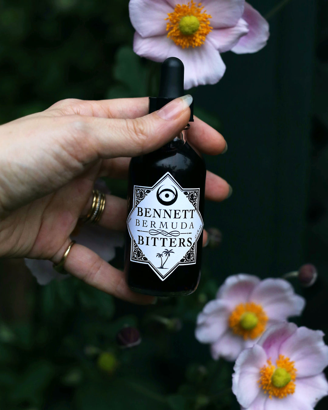 Hand holding a bottle of Tropical Bermuda Bitters by Bennett Bitters in front of pink flowers.
