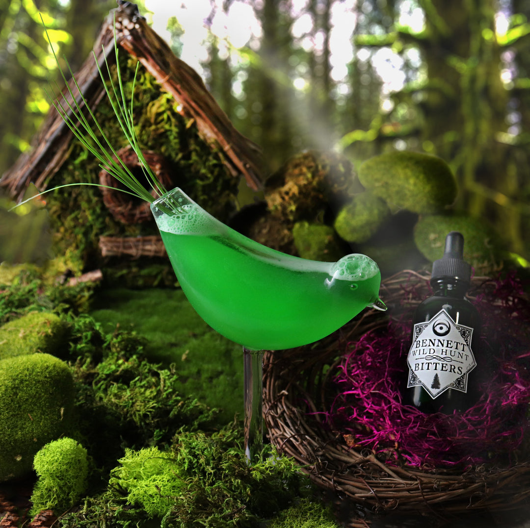 A bottle of Herbal Wild Hunt Bitters by Bennett Bitters inside a birds nest, next to a green cocktail in a bird shaped glass; in a woodland setting.