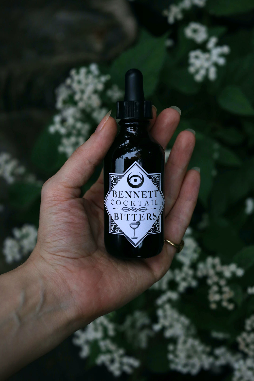 Hand holding a bottle of Old Fashioned Cocktail Bitters by Bennett Bitters in front of white flowers.