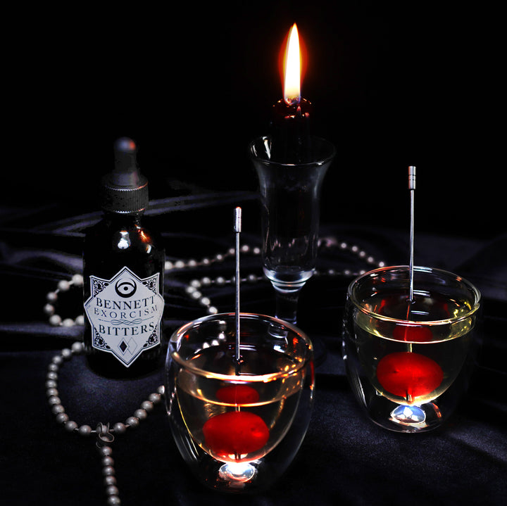 Aromatic Exorcism Bitters bottle by Bennett Bitters, two cocktails with red cherries, and a black candle on black fabric.