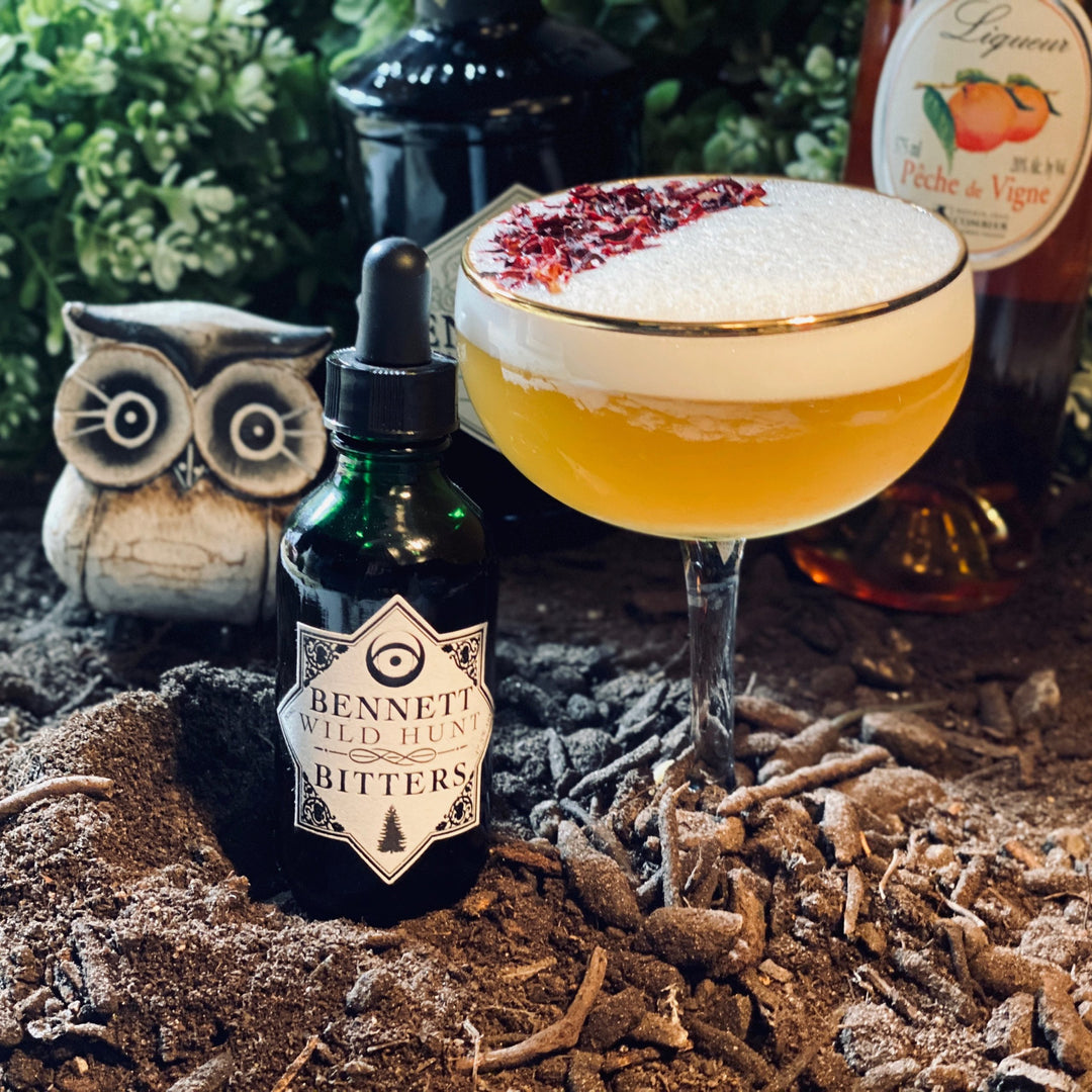 A bottle of Herbal Wild Hunt Bitters by Bennett Bitters next to an apricot cocktail in a forest floor scene.