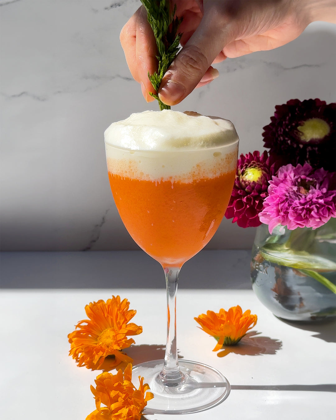 A hand garnishing The Carrot Cake cocktail, a bright orange drink in a Nick and Norah glass topped with white fluffy cream cheese foam.