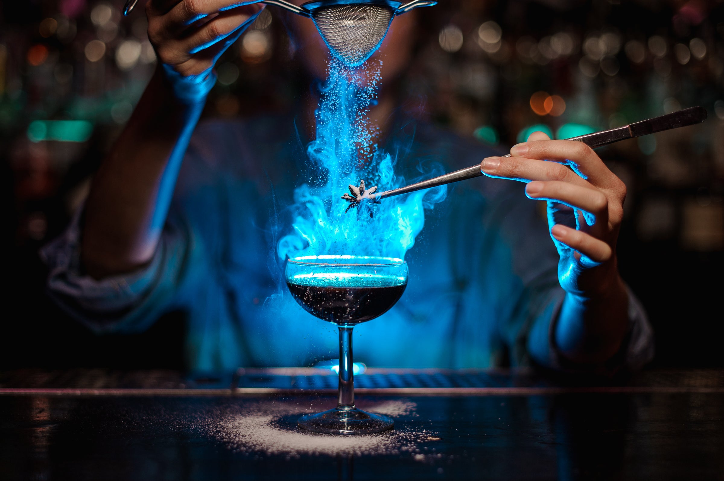 A bartender, in a dark bar lit with blue light, shakes illuminated dust over a cocktail with one hand, while garnishing the drink with a star anise held by tweezers in their other hand.  