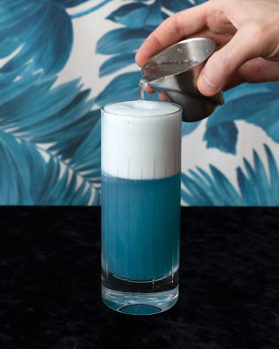 Hand pouring soda water into a blue gin fizz cocktail with tall foamy head.