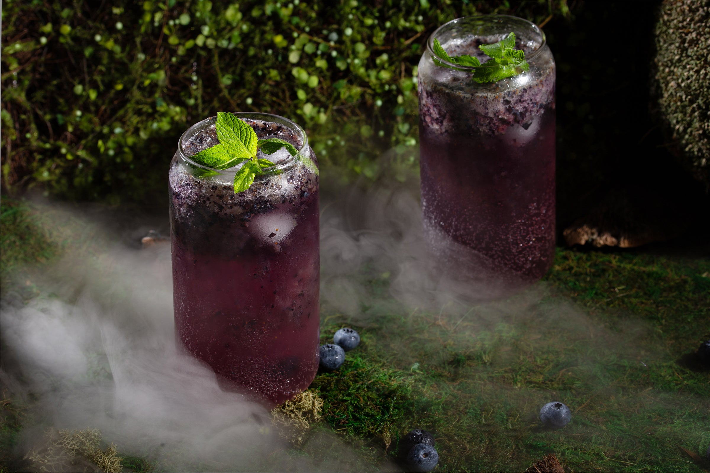 Blue and purple cocktail with mint garnish and blueberries on a misty and mossy forest floor.