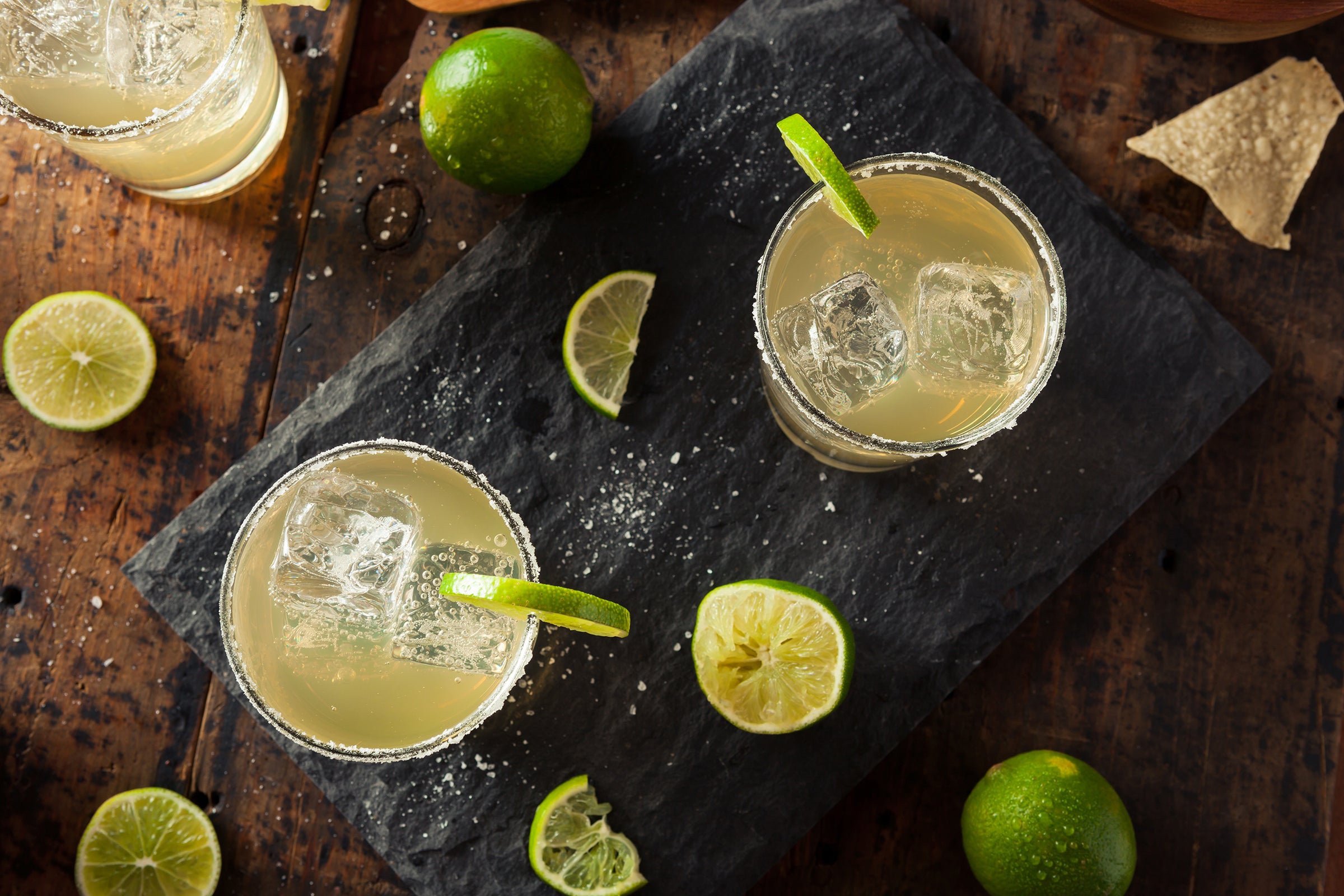 Margaritas and limes scattered about a grey slate board on a dark wood table.