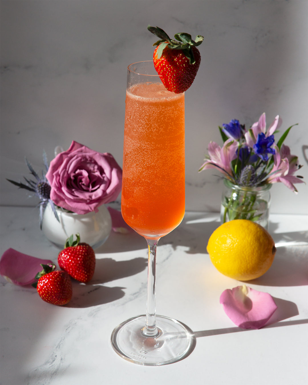 An Ideal Husband strawberry Champagne margarita in a Champagne flute with a strawberry garnish in a scene with flowers, strawberries, and a lemon.