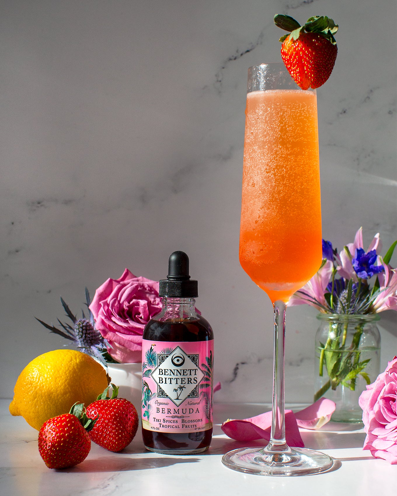 An Ideal Husband strawberry Champagne margarita in a Champagne flute next to a bottle of tropical Bermuda Bitters, a lemon, strawberries, and flowers.
