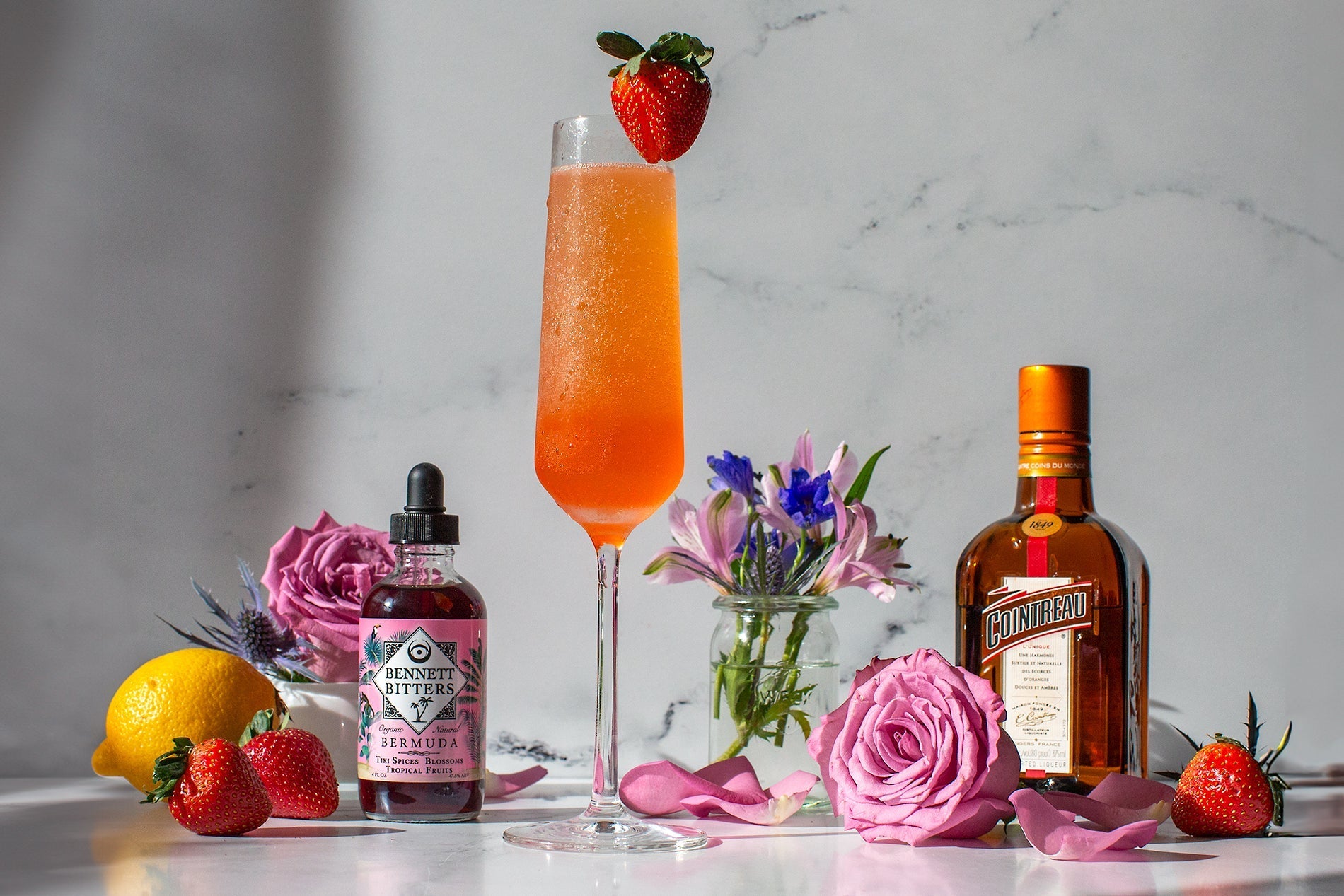 The Ideal Husband strawberry Champagne margarita in a white marble setting  with flowers, a bottle of Cointreau, a bottle of Bermuda Bitters, strawberries, and a lemon.