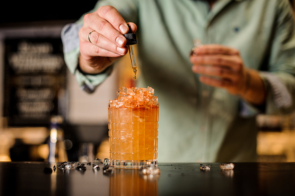 Hand dripping bitters into an orange colored cocktail with ice.